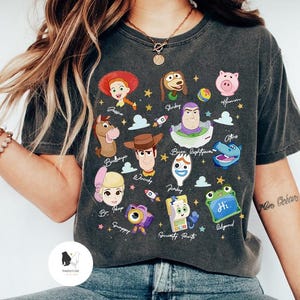 May include: Dark gray t-shirt featuring colorful cartoon character illustrations from the movie Toy Story. The shirt includes characters like Woody, Buzz Lightyear, and Jessie, with text labels. A fun and playful design.