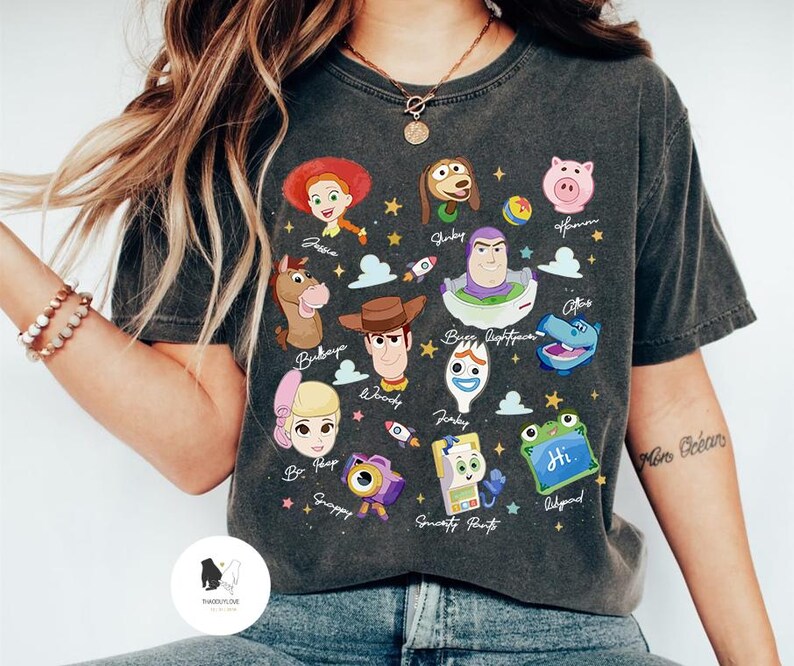 May include: Dark gray t-shirt featuring colorful cartoon character illustrations from the movie Toy Story. The shirt includes characters like Woody, Buzz Lightyear, and Jessie, with text labels. A fun and playful design.