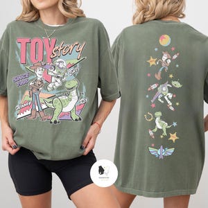 May include: Olive green t-shirt featuring the Toy Story logo and characters Woody, Buzz, and Rex. The back of the shirt displays additional Toy Story characters and space-themed graphics. The text "TOY Story" and "Since 1995" are visible.