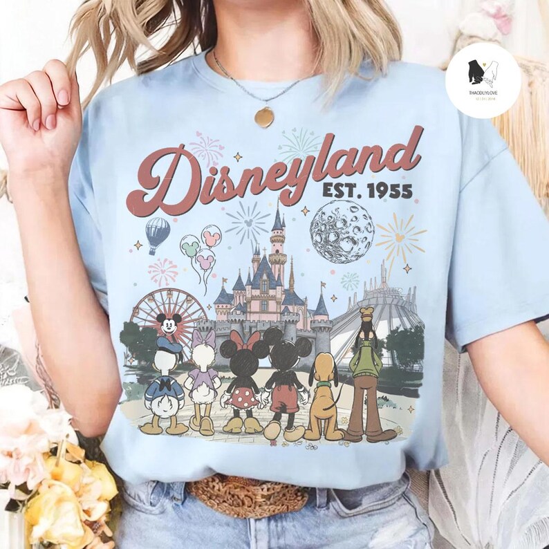 May include: A light blue t-shirt featuring a graphic print of Disneyland with the text "Disneyland EST. 1955". The design includes a castle, a moon, fireworks, and cartoon characters including Mickey Mouse, Minnie Mouse, Donald Duck, and Goofy.
