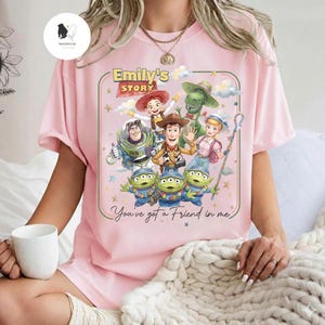 May include: Pink t-shirt featuring a colorful graphic of characters from the animated movie Toy Story. The design includes the text "Emily's Story" and "You've got a Friend in me."