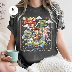 May include: Dark gray t-shirt featuring a colorful graphic of characters from "Toy Story." The design includes Woody, Buzz Lightyear, Jessie, Rex, and the Little Green Men, with the text "Emily's Story" and "You've got a Friend in me."