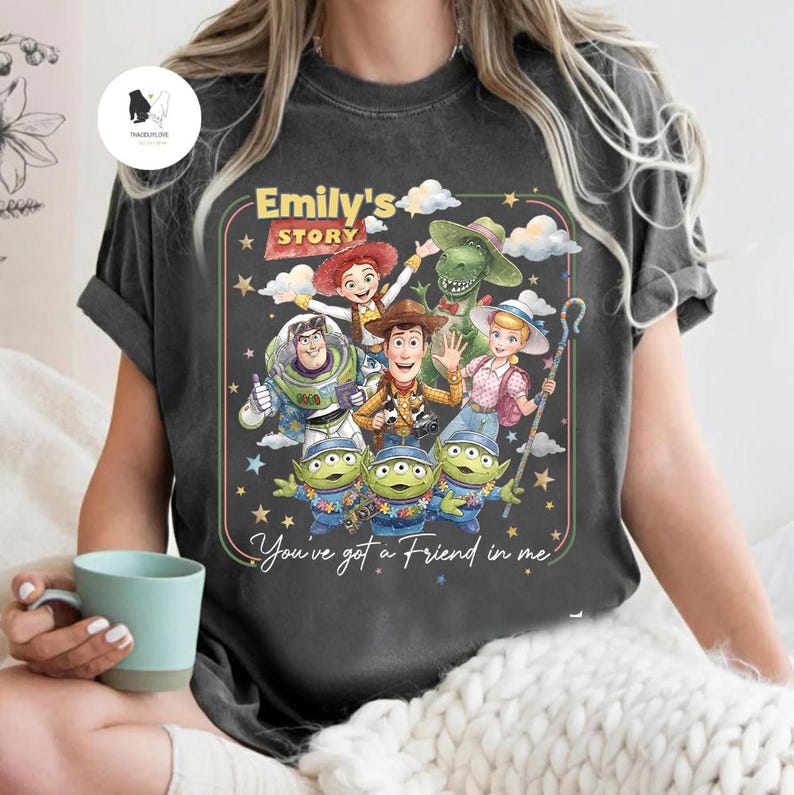 May include: Dark gray t-shirt featuring a colorful graphic of characters from "Toy Story." The design includes Woody, Buzz Lightyear, Jessie, Rex, and the Little Green Men, with the text "Emily's Story" and "You've got a Friend in me."