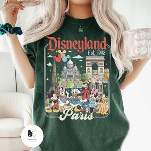 May include: Dark green t-shirt featuring a Disneyland Paris graphic. The design includes iconic landmarks like the Eiffel Tower and Arc de Triomphe, along with Disney characters such as Mickey Mouse, Donald Duck, and Goofy. The text "Disneyland Est. 1992 Paris" is displayed.