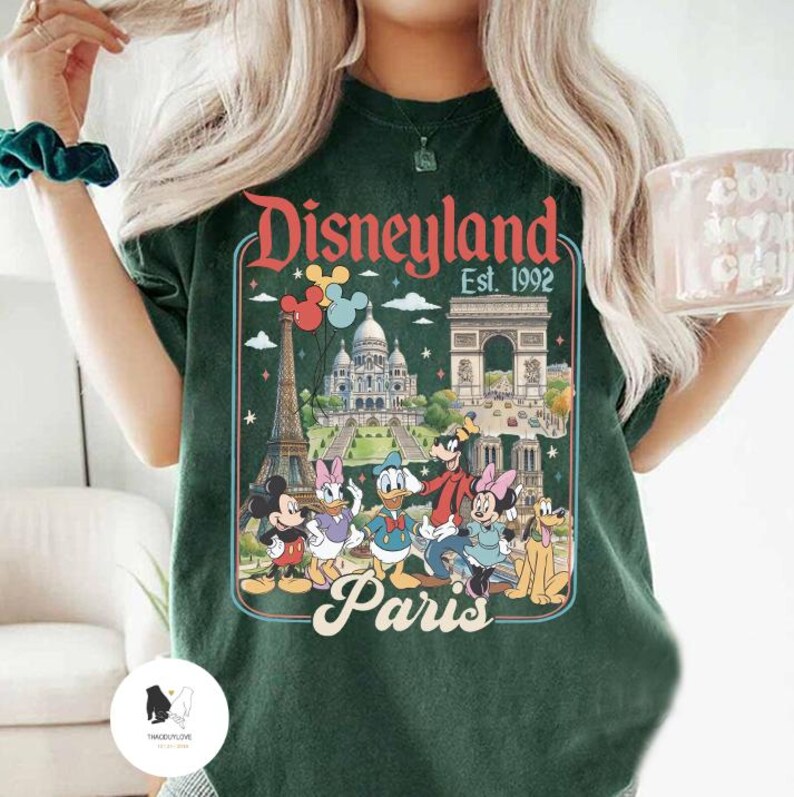 May include: Dark green t-shirt featuring a Disneyland Paris graphic. The design includes iconic landmarks like the Eiffel Tower and Arc de Triomphe, along with Disney characters such as Mickey Mouse, Donald Duck, and Goofy. The text "Disneyland Est. 1992 Paris" is displayed.