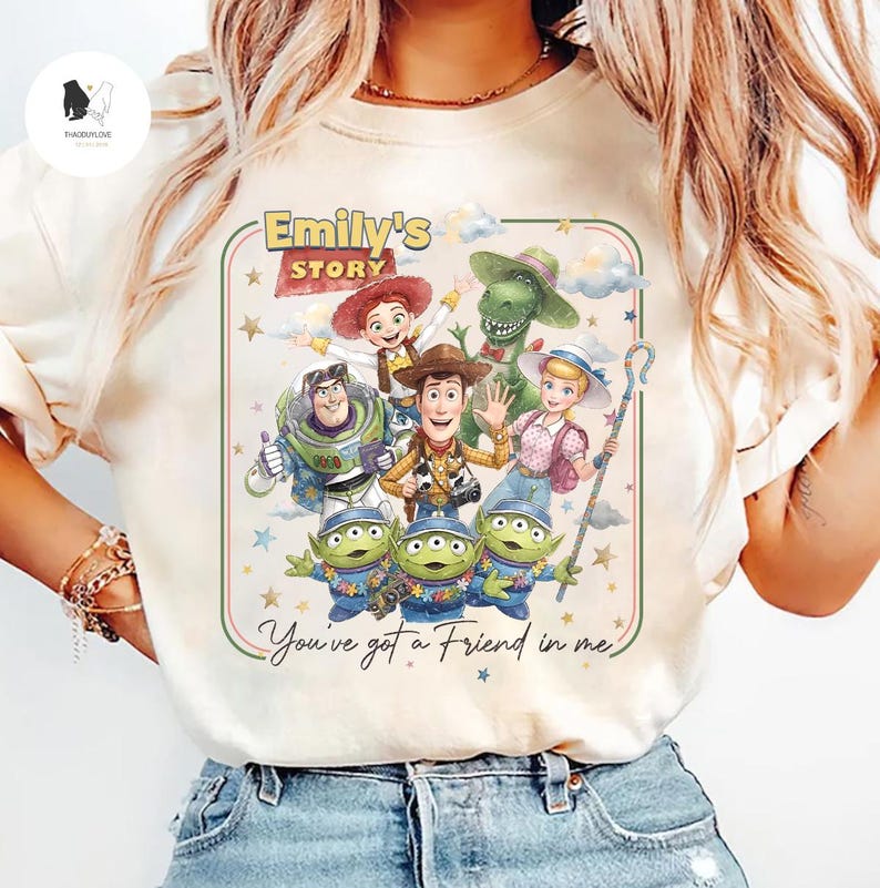 May include: Cream-colored t-shirt featuring a colorful graphic of characters from the animated film "Toy Story." The design includes Woody, Buzz Lightyear, Jessie, Rex, Bo Peep, and the Little Green Men, with the text "Emily's Story" and "You've got a Friend in me."