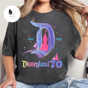 Op de afbeelding: Een zwart T-shirt met een kleurrijke afbeelding van het Disneyland-kasteel en de tekst "Disneyland 70" met de jaren 1955 en 2025.