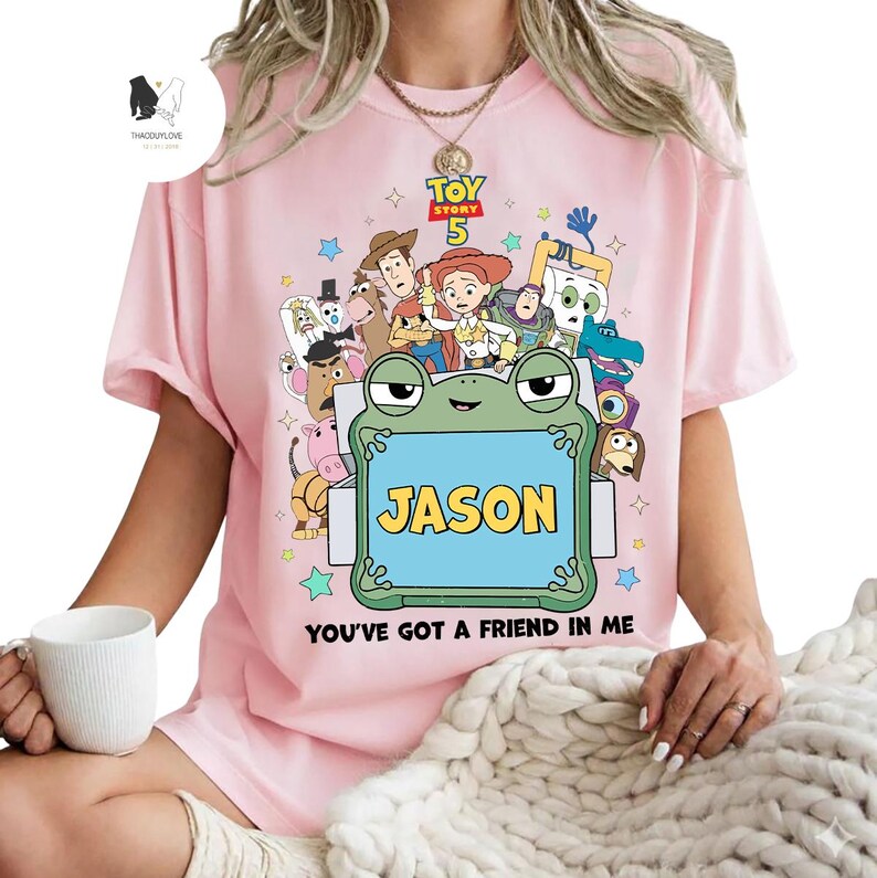 May include: A light pink t-shirt featuring a colorful graphic of characters from the movie Toy Story 5. The shirt includes the name "JASON" on a blue sign held by a frog, with the phrase "YOU'VE GOT A FRIEND IN ME" below.