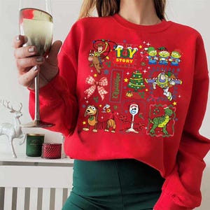 May include: Red sweatshirt featuring a colorful Toy Story Christmas design. The graphic includes Woody, Buzz Lightyear, and other characters, along with a Christmas tree and the words "Toy Story Christmas".