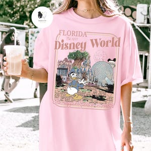 Puede incluir: Camiseta rosa claro con un dise&ntilde;o gr&aacute;fico de Donald Duck frente a los monumentos de Disney World. El dise&ntilde;o incluye el texto "FLORIDA Disney World Est. 1971" y "The happiest place on earth" en estilo vintage.