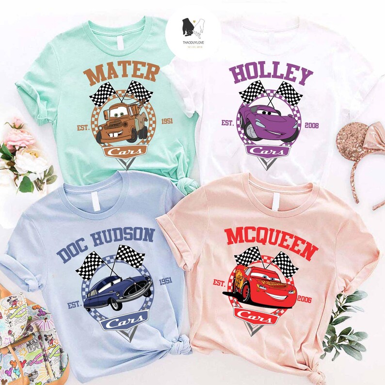 May include: Four t-shirts featuring different Cars characters. The shirts are light blue, pink, white and light green. Each shirt has a chequered flag design and the character's name. The text on the shirts reads "Mater", "Holley", "Doc Hudson", and "McQueen".