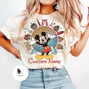 May include: Cream-colored t-shirt featuring a colorful graphic of Mickey Mouse with his arms outstretched, surrounded by landmarks. The text "Custom Name" is below the graphic. The shirt is a casual style.