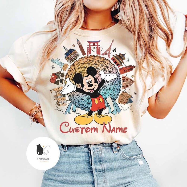 May include: Cream-colored t-shirt featuring a colorful graphic of Mickey Mouse with his arms outstretched, surrounded by landmarks. The text "Custom Name" is below the graphic. The shirt is a casual style.