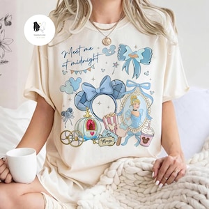 May include: Cream-colored t-shirt featuring a Cinderella-themed design. The design includes a blue bow, a carriage, and the text "Meet me at midnight." A portrait of Cinderella is also included in the design.