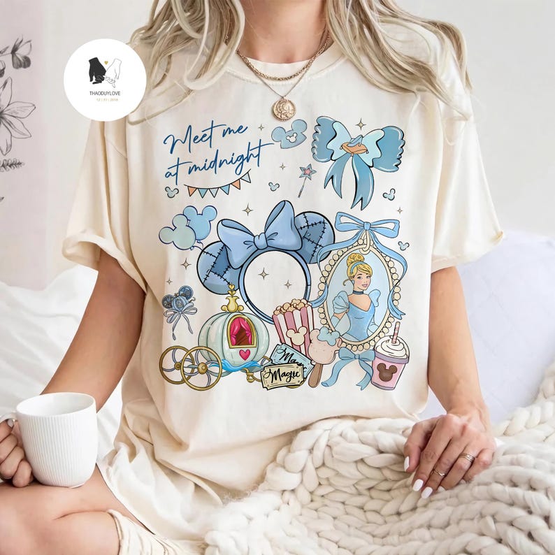 May include: Cream-colored t-shirt featuring a Cinderella-themed design. The design includes a blue bow, a carriage, and the text "Meet me at midnight." A portrait of Cinderella is also included in the design.