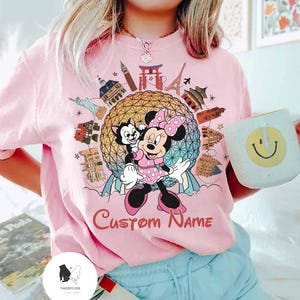 May include: Pink t-shirt featuring Minnie Mouse and Figaro in front of a globe with world landmarks. The shirt has the text "Custom Name" in a cursive font. A white mug with a smiley face is held in the hand.