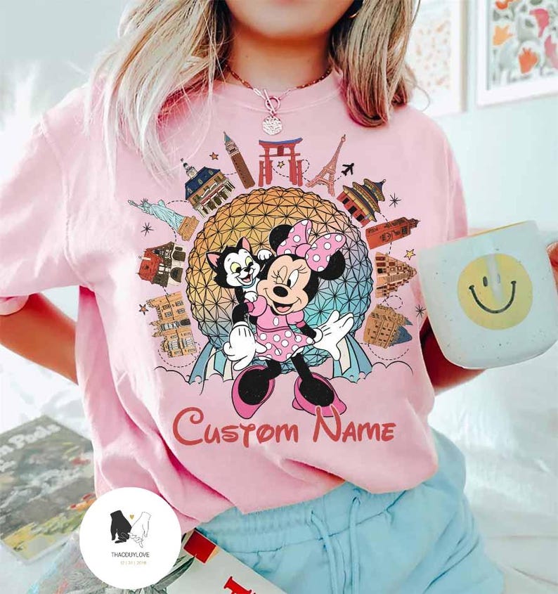 May include: Pink t-shirt featuring Minnie Mouse and Figaro in front of a globe with world landmarks. The shirt has the text "Custom Name" in a cursive font. A white mug with a smiley face is held in the hand.