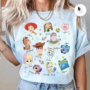May include: Light blue t-shirt featuring colorful cartoon character illustrations from the movie Toy Story. Characters include Woody, Buzz Lightyear, Jessie, and others. The shirt has a crew neck and short sleeves.
