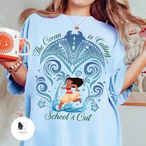 Op de afbeelding: Een lichtblauw t-shirt met een afbeelding van een personage dat op een strand rent met palmbomen en oceaangolven. De tekst The Ocean is Calling en School's Out staat in donkergroen schrift. Een gestileerde mantarog staat boven het personage.