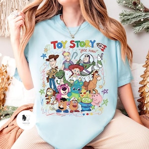 May include: A light blue t-shirt featuring a colorful graphic print of various Toy Story characters including Woody, Buzz Lightyear, Jessie, and others, with the text "TOY STORY" and "yee haw" surrounded by scribbled star and circle doodles.