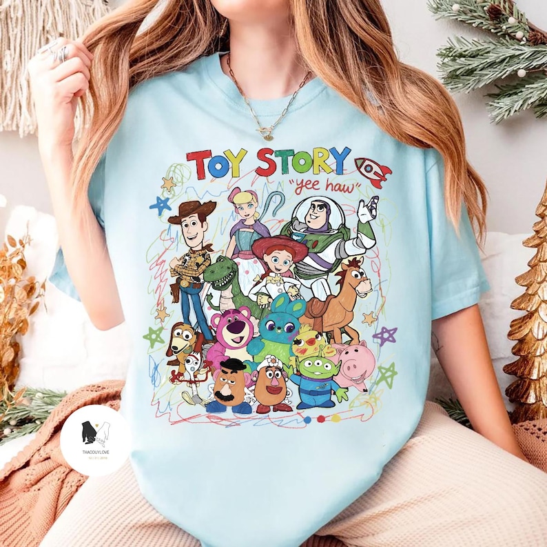 May include: A light blue t-shirt featuring a colorful graphic print of various Toy Story characters including Woody, Buzz Lightyear, Jessie, and others, with the text "TOY STORY" and "yee haw" surrounded by scribbled star and circle doodles.