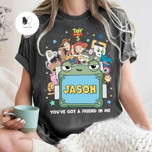 May include: Dark gray t-shirt featuring characters from Toy Story 5. The design includes a frog-shaped frame with the name "JASON" on a blue background. The text "YOU'VE GOT A FRIEND IN ME" is below the frog.