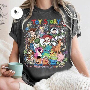 May include: A charcoal gray short-sleeve t-shirt featuring a colorful graphic print of Toy Story characters including Woody, Buzz Lightyear, Jessie, and others. The design includes the text "TOY STORY" and "yee haw" with scribbled line art.