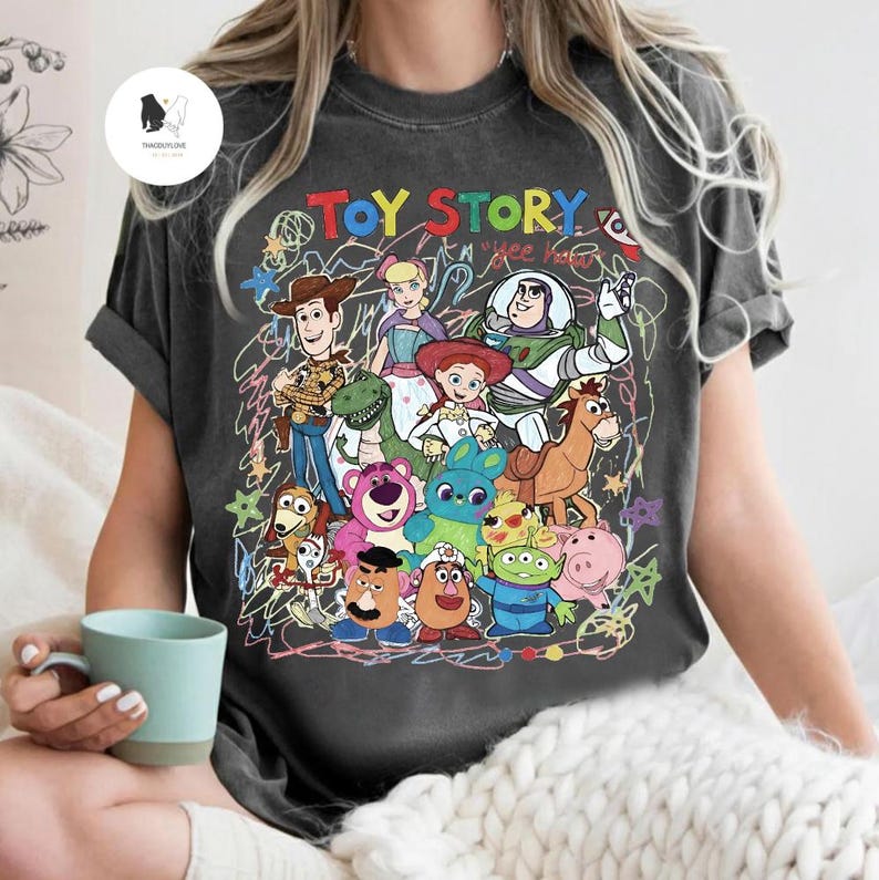 May include: A charcoal gray short-sleeve t-shirt featuring a colorful graphic print of Toy Story characters including Woody, Buzz Lightyear, Jessie, and others. The design includes the text "TOY STORY" and "yee haw" with scribbled line art.