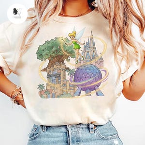 Op de afbeelding: Cr&egrave;mekleurig T-shirt met een kleurrijke illustratie van Tinkerbell met een toverstaf, het Hollywood Tower Hotel, een kasteel en een geometrische bol. Het shirt heeft een vintage, licht vervaagde uitstraling.