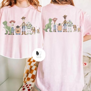 Peut inclure: T-shirt rose clair avec un dessin anim&eacute; de personnages d'un film d'animation populaire. Le devant et le dos du t-shirt pr&eacute;sentent le m&ecirc;me motif, avec un dinosaure, une cow-girl, une t&ecirc;te de pomme de terre, un cow-boy, un ranger de l'espace et un chien.