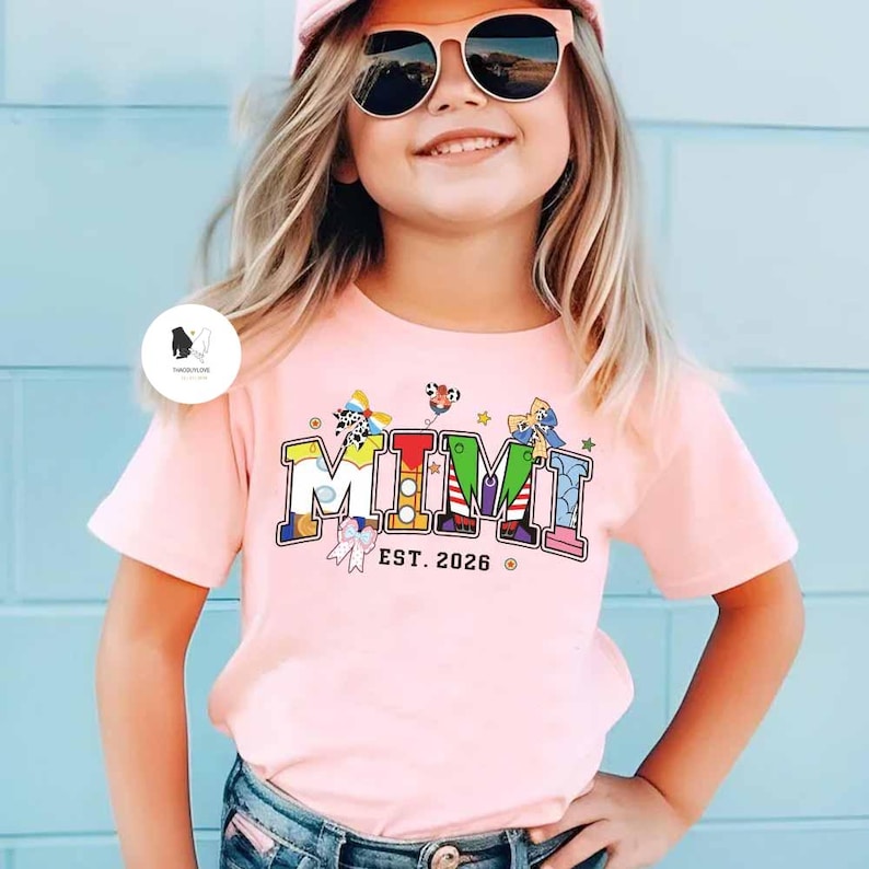 May include: A light pink t-shirt with the word "MIMI" in colourful block letters. The shirt also features the text "EST. 2026" in a smaller font. The shirt is worn by a young person wearing sunglasses.