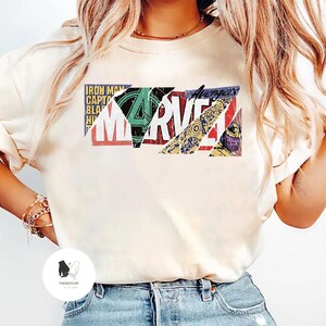 Op de afbeelding: Cr&egrave;mekleurige sweatshirt met een kleurrijk Marvel-logo-ontwerp. Het logo is samengesteld uit verschillende elementen in stripstijl, waaronder het Avengers-logo en tekst als "Iron Man" en "Captain America". De trui is een casual stijl.