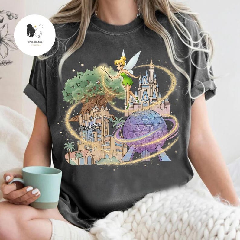 Op de afbeelding: Donkergrijze t-shirt met een kleurrijke afbeelding van Tinkerbell met een toverstaf, omringd door iconische Disney-park bezienswaardigheden zoals het Hollywood Tower Hotel, Spaceship Earth en Cinderella Castle. Het ontwerp is omlijnd met gouden glitters.
