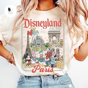 May include: Cream-colored t-shirt featuring a Disneyland Paris graphic. The design includes the Eiffel Tower, Arc de Triomphe, and Disney characters like Mickey Mouse, Donald Duck, and Goofy. The text reads "Disneyland Paris Est. 1992."