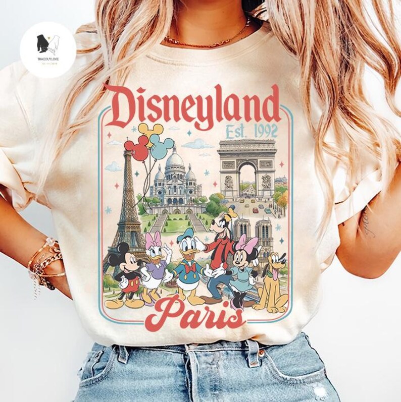 May include: Cream-colored t-shirt featuring a Disneyland Paris graphic. The design includes the Eiffel Tower, Arc de Triomphe, and Disney characters like Mickey Mouse, Donald Duck, and Goofy. The text reads "Disneyland Paris Est. 1992."