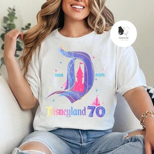 Op de afbeelding: Een wit T-shirt met een kleurrijke afbeelding van het Disneyland-kasteel en de tekst "Disneyland 70" met de jaren 1955 en 2025 aan weerszijden van het kasteel.