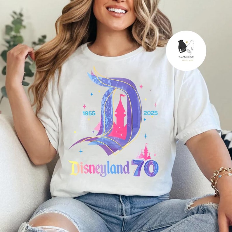 Op de afbeelding: Een wit T-shirt met een kleurrijke afbeelding van het Disneyland-kasteel en de tekst "Disneyland 70" met de jaren 1955 en 2025 aan weerszijden van het kasteel.