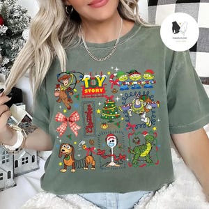 May include: Sage green t-shirt featuring a colorful graphic of Toy Story characters and the text "Toy Story Christmas". The design includes Woody, Buzz Lightyear, aliens, and other characters in a festive holiday theme.