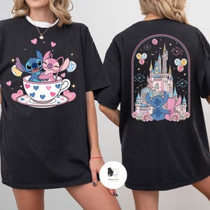 May include: Black t-shirt featuring a cartoon design of Stitch and Angel. The front shows them in a teacup with hearts, while the back displays a castle scene with fireworks and balloons. A cute and colorful design.