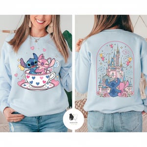 May include: Light blue sweatshirt featuring a cartoon illustration of Stitch and Angel in a teacup with hearts. The back of the sweatshirt has a castle design with Stitch and Angel. The sweatshirt has a crew neck and long sleeves.