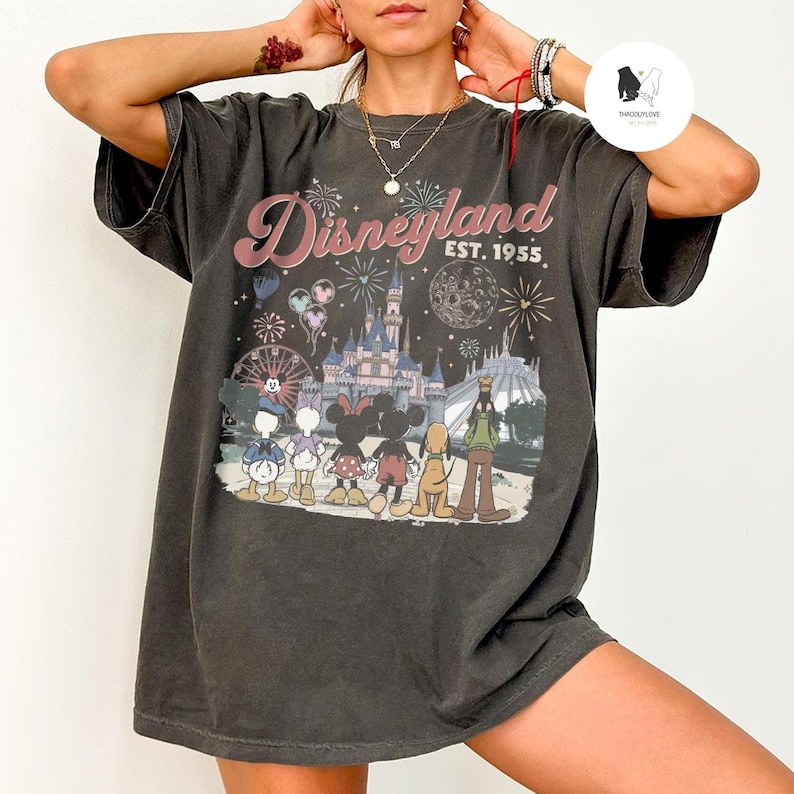 May include: A charcoal grey short-sleeve t-shirt featuring a graphic print of Disney characters standing before a castle. The text reads Disneyland Est. 1955. The design includes fireworks, a moon, and a ferris wheel in a muted colour palette.