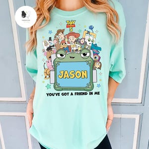 May include: A light blue t-shirt featuring a colorful graphic of characters from the movie Toy Story 5. The design includes the text "JASON" in a blue frame and the phrase "YOU'VE GOT A FRIEND IN ME".