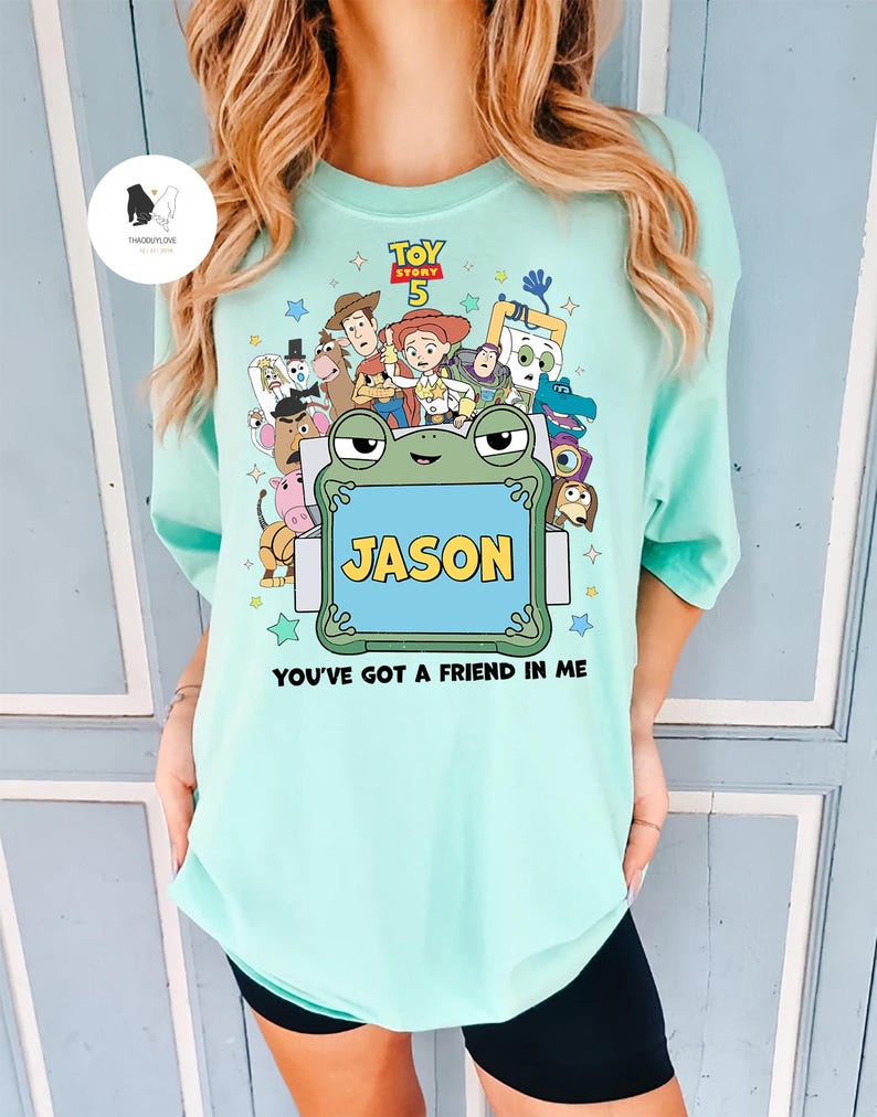 May include: A light blue t-shirt featuring a colorful graphic of characters from the movie Toy Story 5. The design includes the text "JASON" in a blue frame and the phrase "YOU'VE GOT A FRIEND IN ME".
