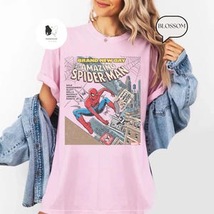 Puede incluir: Camiseta rosa claro con un dise&ntilde;o de portada de c&oacute;mic de Spider-Man. El gr&aacute;fico incluye el texto "BRAND NEW DAY" y "The Amazing Spider-Man". El superh&eacute;roe est&aacute; representado con su cl&aacute;sico traje rojo y azul.