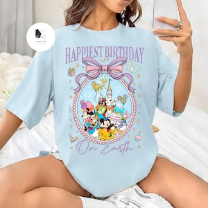 May include: Light blue t-shirt with the text "Happiest Birthday On Earth" and a colorful graphic featuring Disney characters, a castle, and birthday decorations. The design includes Mickey Mouse, Minnie Mouse, and Donald Duck.