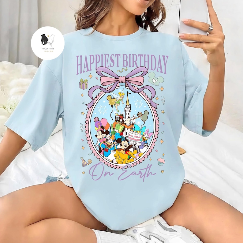 May include: Light blue t-shirt with the text "Happiest Birthday On Earth" and a colorful graphic featuring Disney characters, a castle, and birthday decorations. The design includes Mickey Mouse, Minnie Mouse, and Donald Duck.