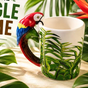 Könnte beinhalten: Ein weißer Keramikbecher mit grünem Tropenblattmuster und einem Henkel in Form eines bunten Ara-Papageis mit roten, blauen und gelben Federn. Der Text BRING THE JUNGLE HOME ist in grünen und braunen Buchstaben über dem Becher aufgedruckt.