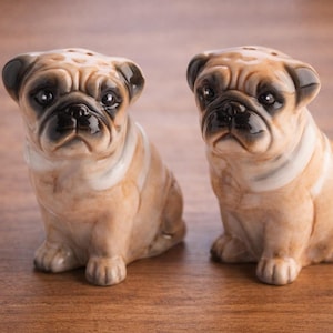 May include: Two ceramic pug dog figurines. The salt and pepper shakers are tan with black accents and white collars. The pug dogs are sitting on a wooden surface. The pug dogs have a detailed, realistic appearance.