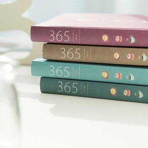 May include: Four stacked daily planners in shades of mauve, brown, teal, and dark teal. The spines display the number "365" and small food and drink illustrations.