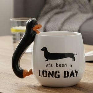 May include: White ceramic mug featuring a black dachshund silhouette and the text "it's been a LONG DAY". The handle is a dachshund dog with black and orange details. A novelty item for dog enthusiasts.